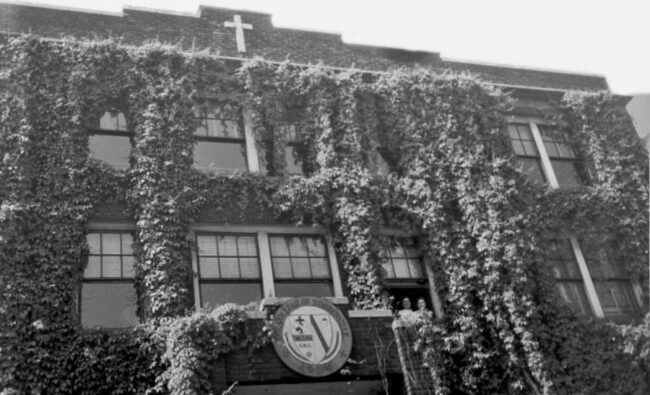 Black and white photo of a building covered in vines with a cross on the top and the Franciscan University seal over the doorway.