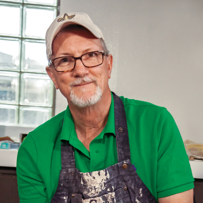 A man in a baseball hat, glasses, green shirt, and clay-covered overalls, sits at a potter's wheel making a bowl.
