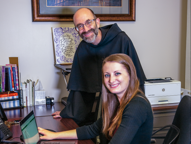 Friar stands while a woman sits at a desk at a laptop in an office.