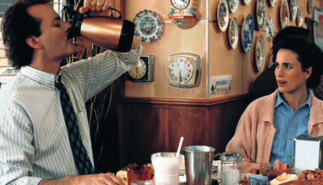 Bill Murray in a dress shirt and tie drinks from the coffee pot as Andie MacDowell looks at him while they sit in a restaurant in the movie Groundhog Day.