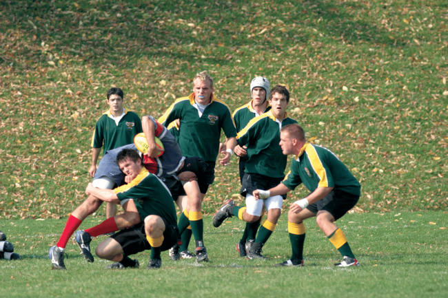 Men in green and yellow rugby uniforms tackle each other in a field.