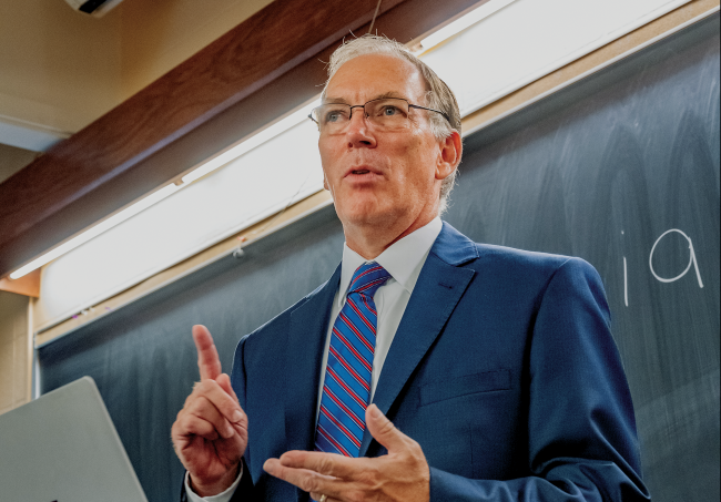 Man in a suit, tie, and glasses, lectures with a computer in front of a chalkboard.