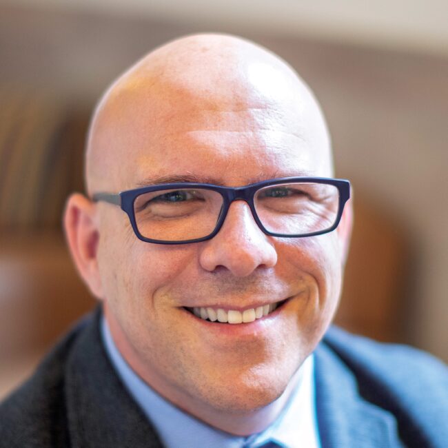 Headshot of Dr. Dan Dentino in a suit, tie, and glasses with a blurred background.