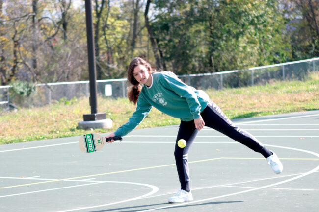 A woman in athletic attire swings at a tennis ball with a paddle on an outdoor tennis court.