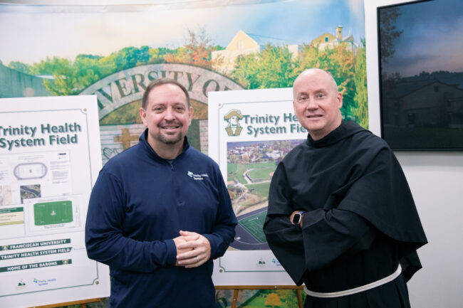 Fr. Dave Pivonka, TOR, in a friar habit, and a man in a Trinity Health Systems shirt, stand in front of a display for an athletic field at Franciscan University.