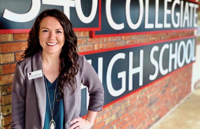 Kimberly Camosse, in formal attire and a name tag, stands in front of a brick wall sign for a high school.
