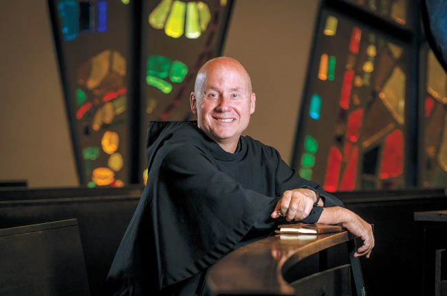 Fr. Dave Pivonka, TOR, in a friar habit, kneels and smiles in a pew in Christ the King Chapel.