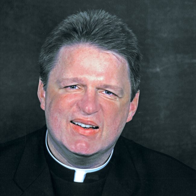 Headshot of Father Stu in priestly cassock in front of a gray background.