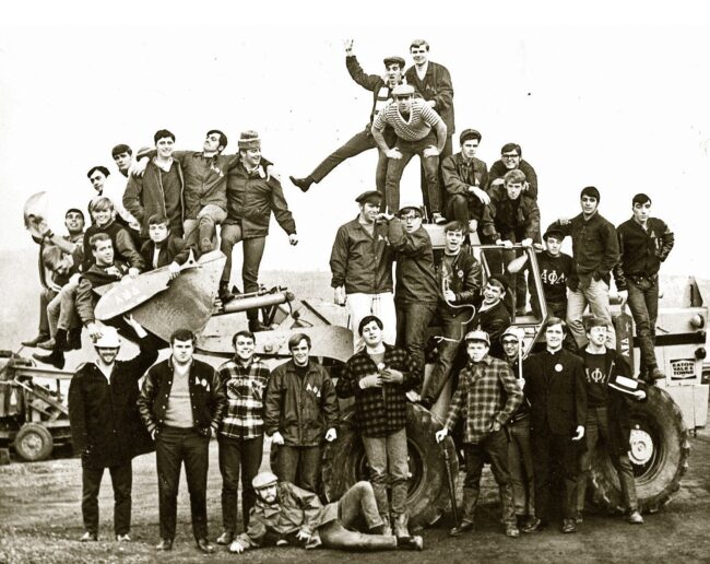Over 30 male students wearing fraternity gear pose precariously atop and alongside a front loader in a black and white photo.