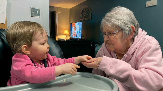 Older woman holds the hand of a young girl who is sitting in a high chair.