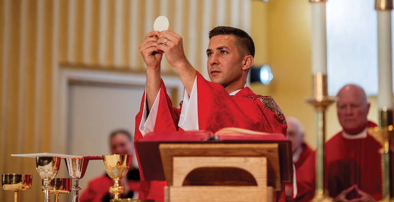 A priest in red priestly robes holds up the Eucharist in Christ the King Chapel
