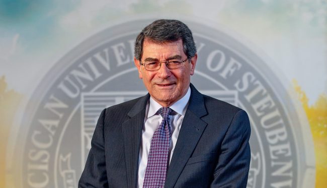 Dr. Michael Healy, in a suit, tie, and glasses, in front of a backdrop with the Franciscan Seal.