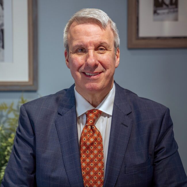 Richard Saxon in a navy suit sitting at his desk with picture frames in the background.