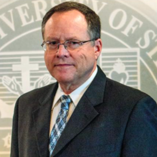 A headshot of Dr. Frank Hermann, in a suit, tie, and glasses, in front of a backdrop with the Franciscan University seal.