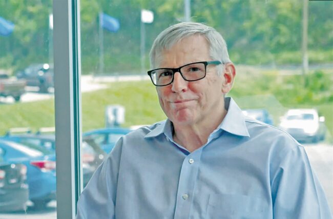 Man in a dress shirt and glasses in front of a glass window looking onto a car lot