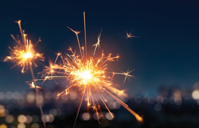 Two sparklers at night in front of a blurred background.