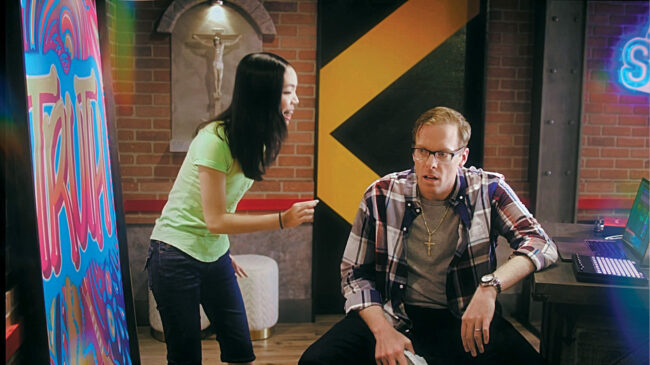 Man in a flannel sits at desk in a colorful room with a brick wall while a woman in a green shirt talks to him.