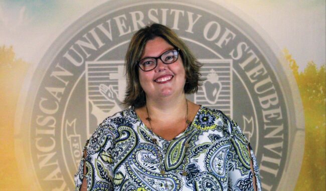 Headshot of Dr. Ann Dulany, in glasses and formal attire, in front of a backdrop with the Franciscan University Seal