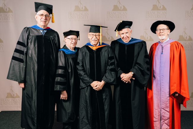A group of five older men in graduation robes in front of a background with the Franciscan University logo.