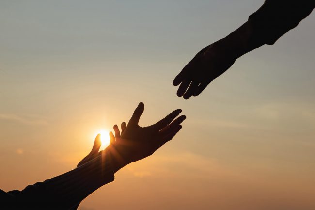 Silhouettes of two hands reaching for each other with the sun setting behind them.