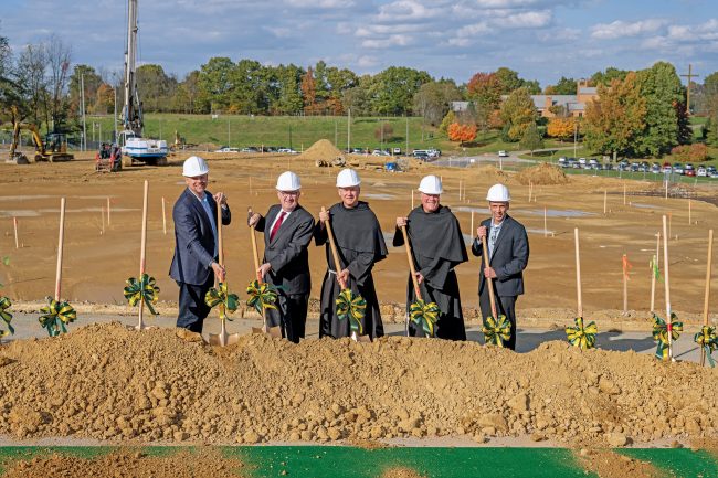 Two friars and three men in suits, all wearing hard hats, hold shovels in front of a pile of dirt.