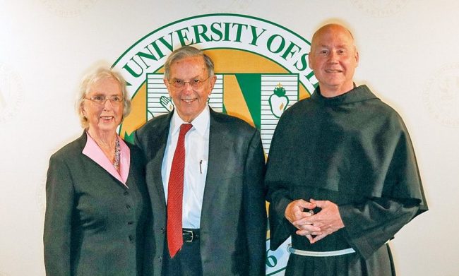 Jane and Nick Healy and Fr. Dave Pivonka standing in front of a university seal.