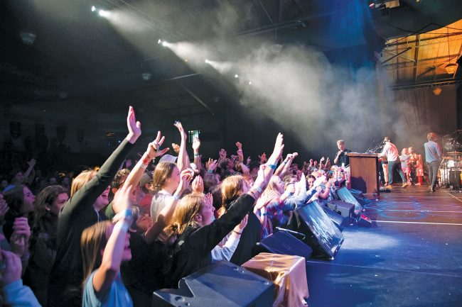 Young people in a concert hall, holding their hands up towards the stage.