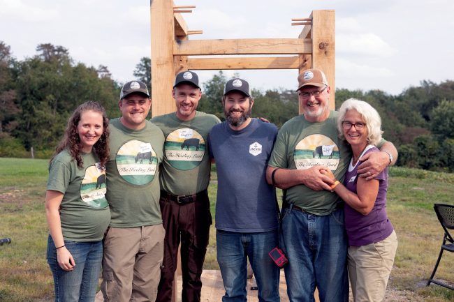 Four men and two women in a group photo outside with a wooden structure behind them.