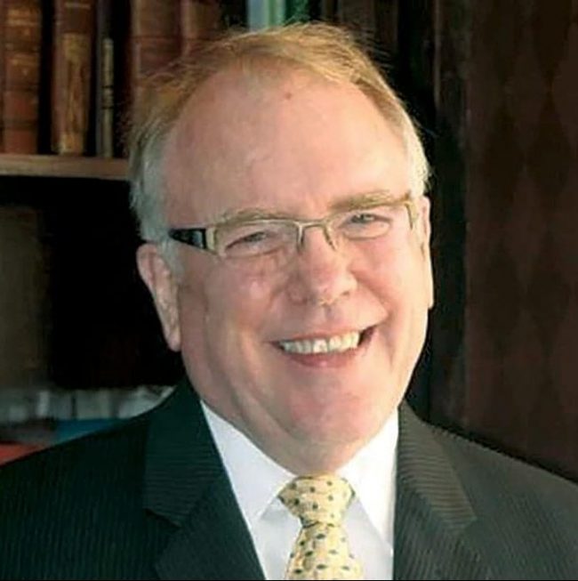 A smiling man in a suit with a bookshelf behind him.
