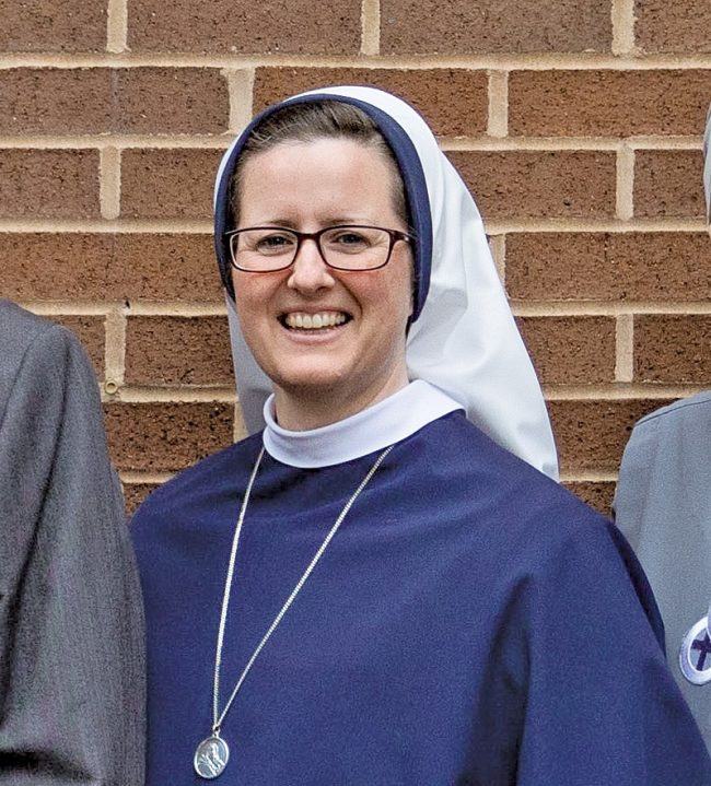 A religious sister in a blue and white habit smiling in front of a brick wall.