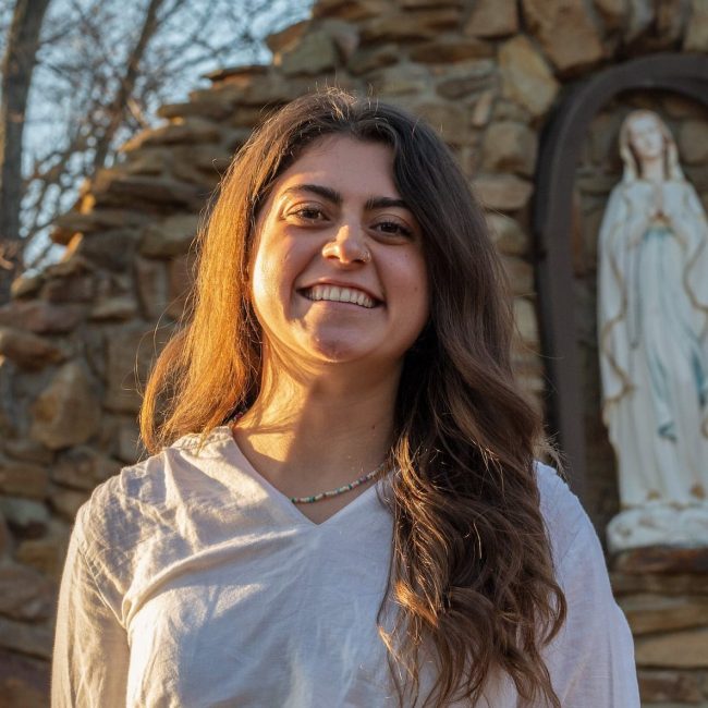 Amelia Brennan in a white shirt smiling in front of grotto with a Mary statue in the background.