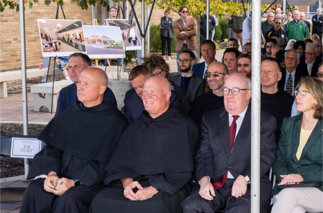 A group of people sitting outside with people standing in the back.