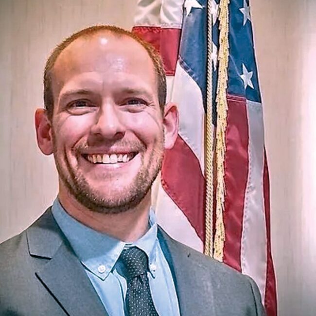 A man in a gray suit smiling in front of a flag.