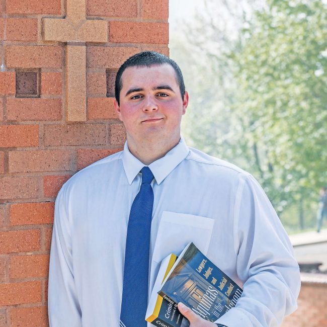 A man in a white button down shirt and tie, holding books and leaning against a brick wall.