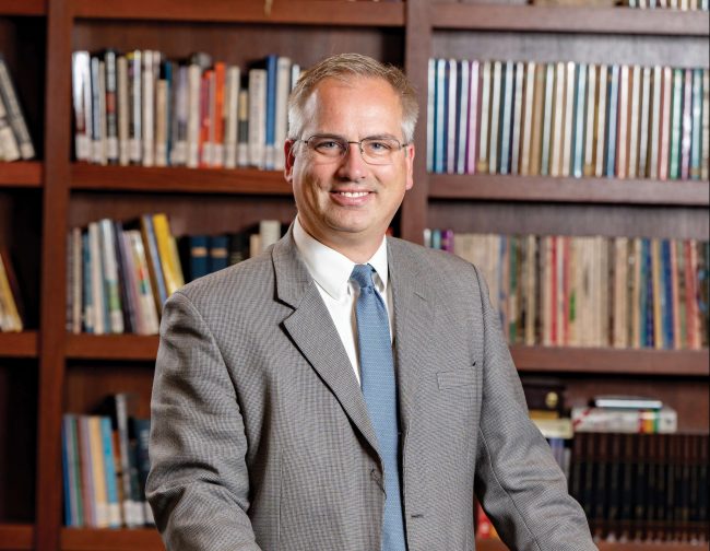 Man in a grey suit in front of book shelves.