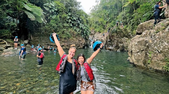 Two women in a lake holding up helmets with other women doing various things in the background.