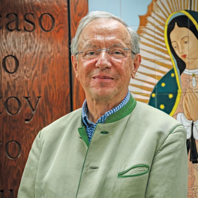 Dr. Michael Waldstein in a green jacket with an image of Our Lady of Guadalupe painted on the wall behind them.