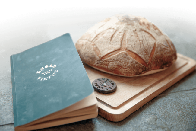 A loaf of bread on a wooden cutting board next to a book that says "Knead Virtue: Gaming 2023" on the cover.