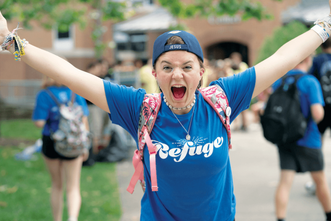 A woman cheering to the camera wearing a blue t-shirt that says "Refuge".