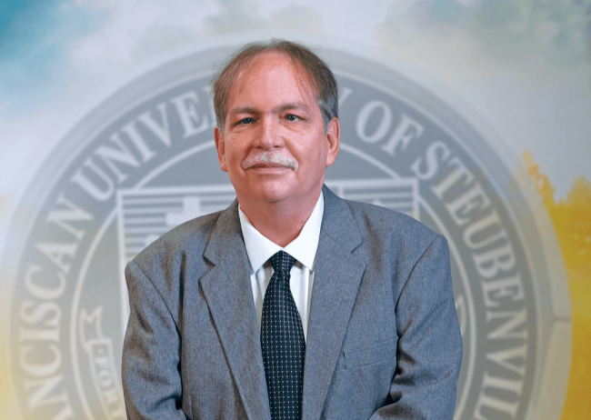 Dr. Tom Kelly in a gray suit standing in front of a university seal background.