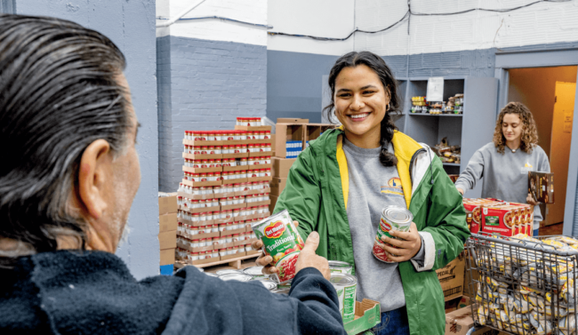 A woman handing a can of tomatos to a man with another woman organizing boxes in the background.