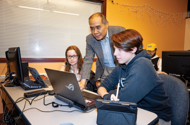 A man in a sport's jacket showing two students something on a laptop.