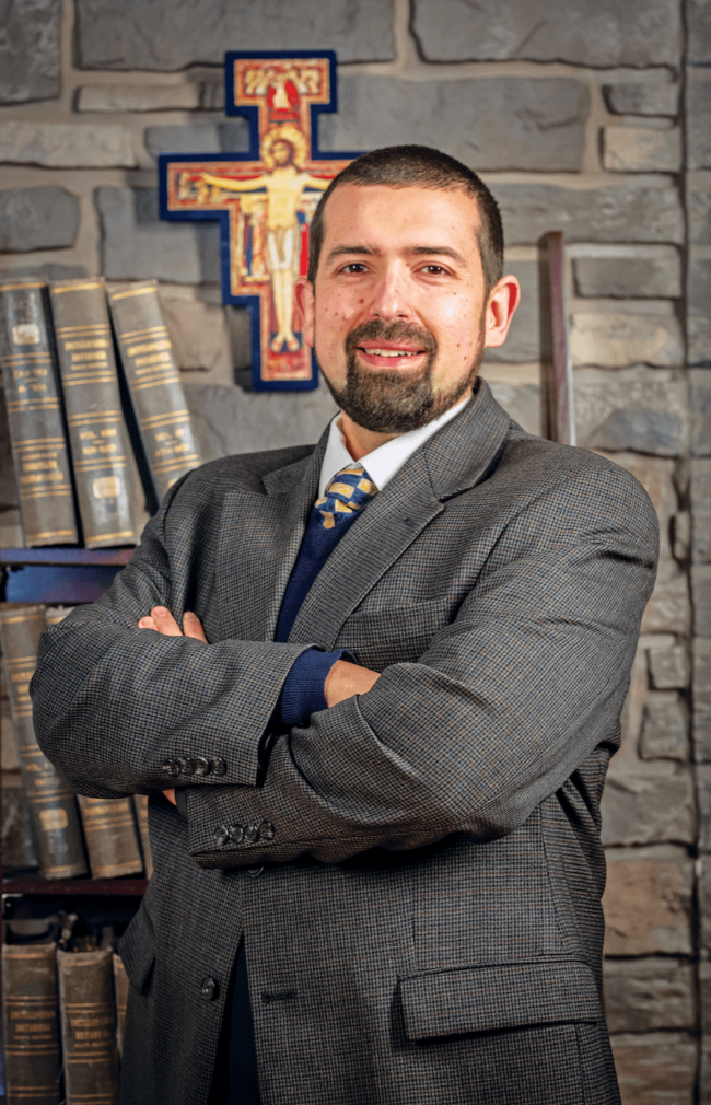 Man in a gray suit in front of a brick wall and books on a shelf and a cross on the wall.
