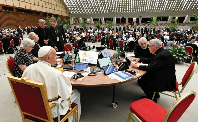 Pope Francis sitting at a round table with a room full of other people at tables behind him.
