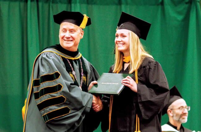 Fr. Dave Pivonka handing a diploma to a woman with a green curtain behind them.