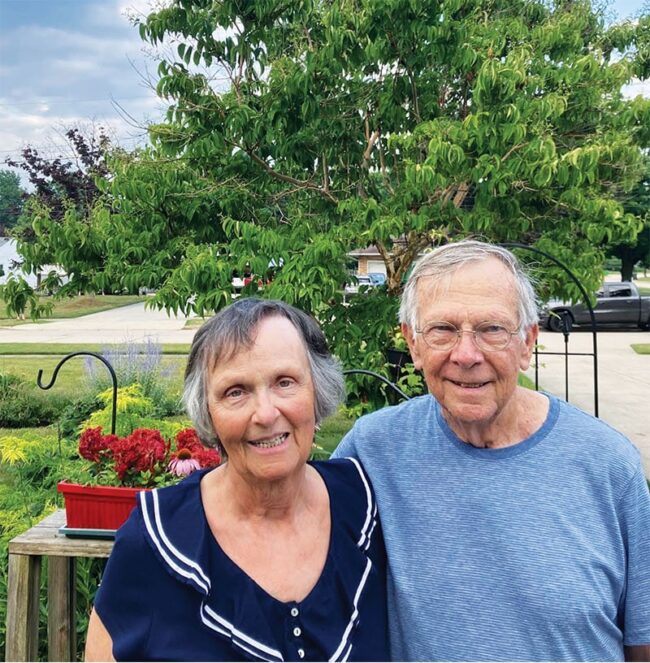 Rosemary (Jereb ’68) and Joe McCormack ’70 standing outside in front of a tree and some flowers.