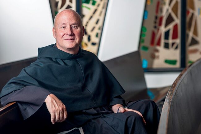 Father Dave Pivonka, TOR, sitting in a church pew with stained glass window.