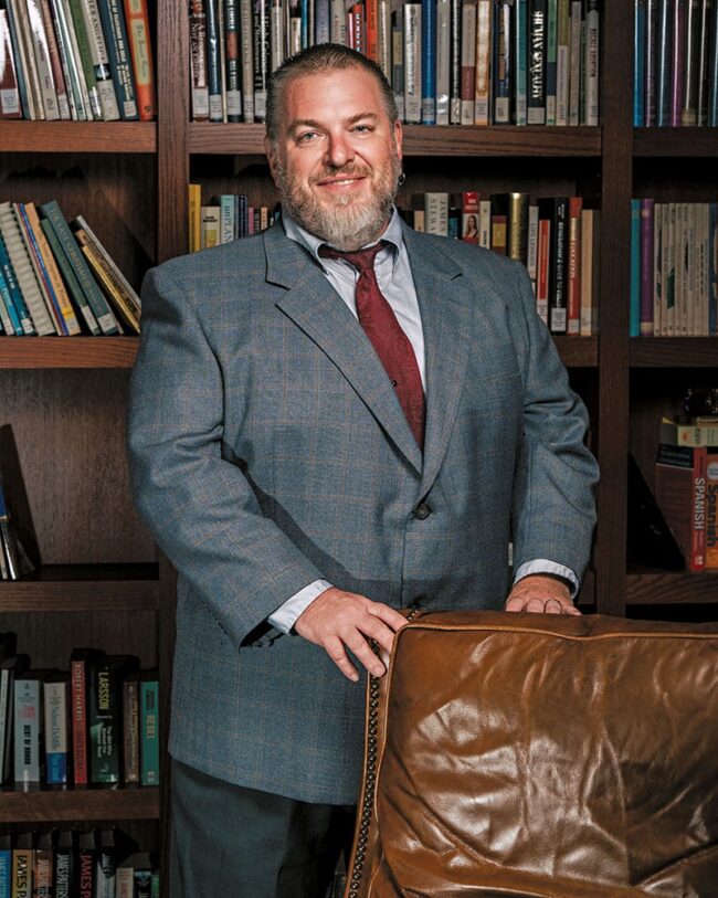 Dr. David Collins in a suit standing in front of a bookshelf.