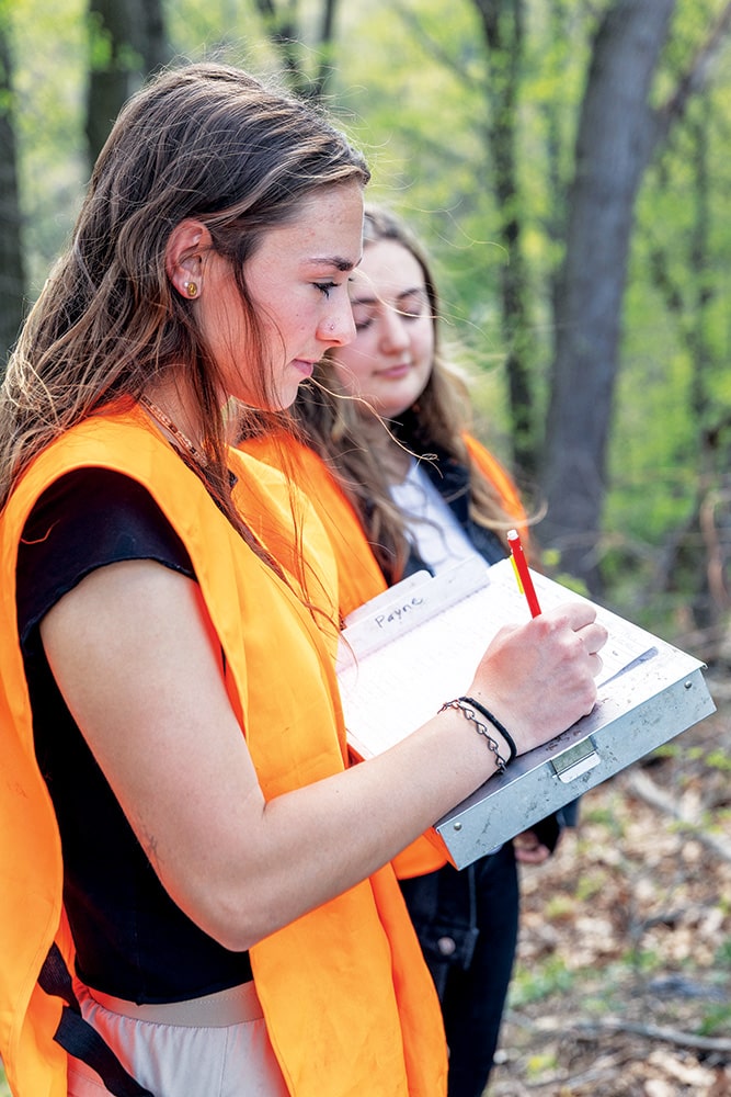 Beth Beigel (foreground) writing notes and Kelly McCarthy standing in a forest. 
