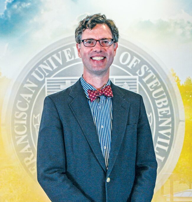 Dr. Alex Plato in a suit and bowtie standing in front of a university seal.